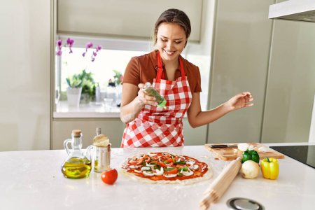 Photo for Young beautiful hispanic woman smiling confident pouring oregano on pizza at the kitchen - Royalty Free Image