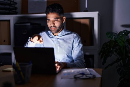 Photo for Hispanic man with beard working at the office with laptop at night pointing with finger to the camera and to you, confident gesture looking serious - Royalty Free Image