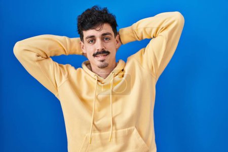 Téléchargez les photos : Homme hispanique debout sur fond bleu relaxant et étirant, bras et mains derrière la tête et le cou souriant heureux - en image libre de droit