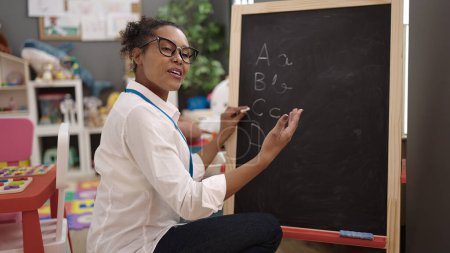 Téléchargez les photos : African american woman teacher smiling confident writing on blackboard at kindergarten - en image libre de droit