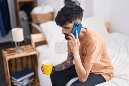 Photo for Young hispanic man talking on smartphone drinking coffee at bedroom - Royalty Free Image