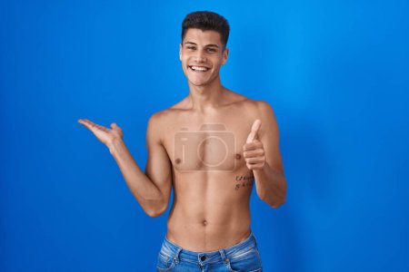 Téléchargez les photos : Jeune homme hispanique debout torse nu sur fond bleu montrant la main de la paume et faisant un geste correct avec les pouces levés, souriant heureux et joyeux - en image libre de droit