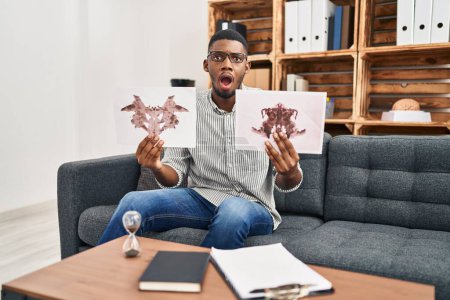 Photo for African american man working with rorschach test at psychology clinic afraid and shocked with surprise and amazed expression, fear and excited face. - Royalty Free Image