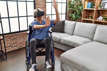 Téléchargez les photos : Jeune homme blond jouant à un jeu vidéo en utilisant des lunettes de réalité virtuelle assis sur un fauteuil roulant à la maison - en image libre de droit