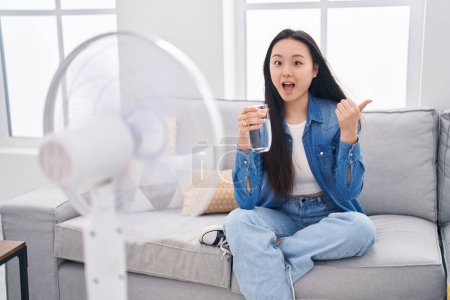 Photo for Young asian woman drinking glass of water enjoying air from fan pointing thumb up to the side smiling happy with open mouth - Royalty Free Image