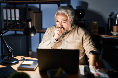 Foto de Hombre de mediana edad con canas trabajando en la oficina por la noche sintiéndose mal y tosiendo como síntoma de resfriado o bronquitis. concepto de atención sanitaria. - Imagen libre de derechos