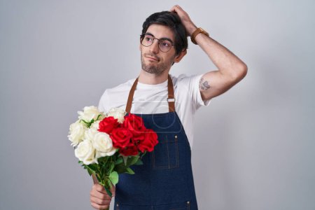 Photo for Young hispanic man holding bouquet of white and red roses confuse and wondering about question. uncertain with doubt, thinking with hand on head. pensive concept. - Royalty Free Image