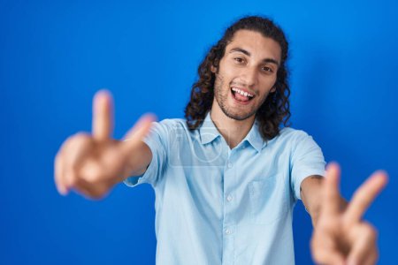 Téléchargez les photos : Jeune homme hispanique debout sur fond bleu souriant avec la langue montrant les doigts des deux mains faisant signe de victoire. numéro deux. - en image libre de droit