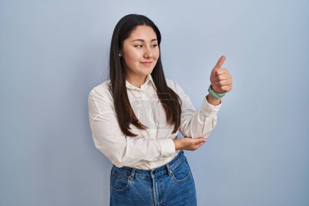 Photo for Young latin woman standing over blue background looking proud, smiling doing thumbs up gesture to the side - Royalty Free Image