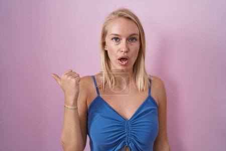 Téléchargez les photos : Young caucasian woman standing over pink background surprised pointing with hand finger to the side, open mouth amazed expression. - en image libre de droit