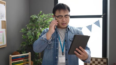 Téléchargez les photos : Young chinese man preschool teacher using touchpad talking on smartphone at kindergarten - en image libre de droit