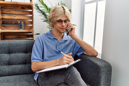 Téléchargez les photos : Young blond man psychologist writing on document talking on smartphone at psychology clinic - en image libre de droit