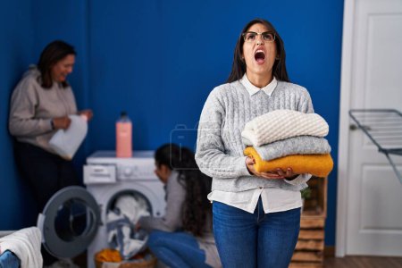 Photo for Three women doing laundry at home angry and mad screaming frustrated and furious, shouting with anger looking up. - Royalty Free Image