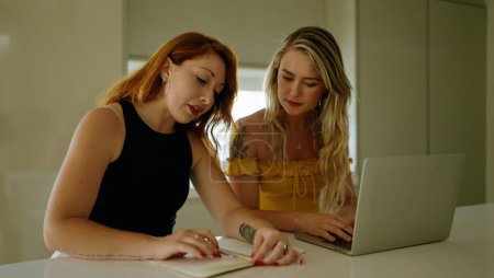 Téléchargez les photos : Two women using laptop writing on notebook sitting on table at kitchen - en image libre de droit