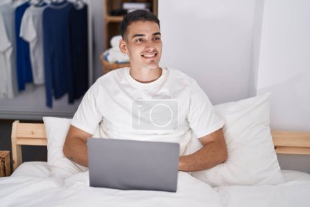 Foto de Hombre hispano joven usando portátil sentado en la cama en el dormitorio - Imagen libre de derechos