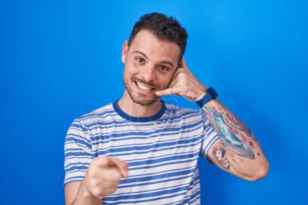 Téléchargez les photos : Young hispanic man standing over blue background smiling doing talking on the telephone gesture and pointing to you. call me. - en image libre de droit