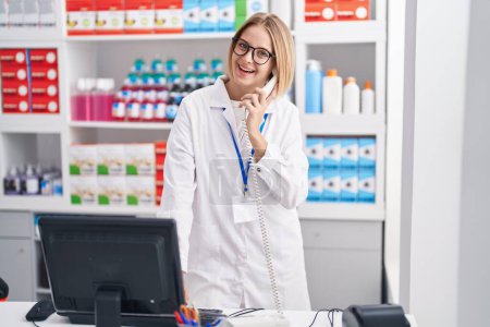Photo for Young blonde woman pharmacist talking on telephone using computer at pharmacy - Royalty Free Image
