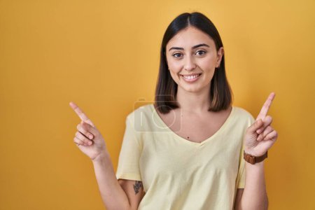 Foto de Chica hispana vistiendo camiseta casual sobre fondo amarillo sonriendo confiado señalando con los dedos a diferentes direcciones. espacio de copia para publicidad - Imagen libre de derechos