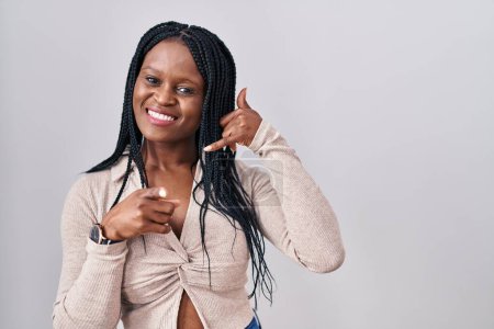 Téléchargez les photos : Femme africaine avec des tresses debout sur fond blanc souriant faire parler au téléphone geste et vous pointant du doigt. Appelle-moi. - en image libre de droit