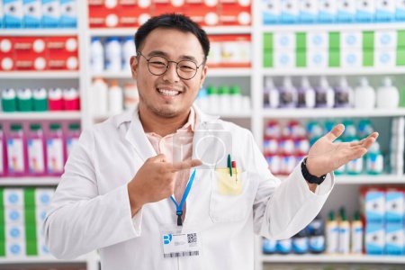 Téléchargez les photos : Jeune homme chinois travaillant à la pharmacie drugstore étonné et souriant à la caméra tout en présentant avec la main et pointant du doigt. - en image libre de droit