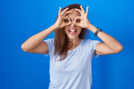 Photo for Brunette woman standing over blue background doing ok gesture like binoculars sticking tongue out, eyes looking through fingers. crazy expression. - Royalty Free Image