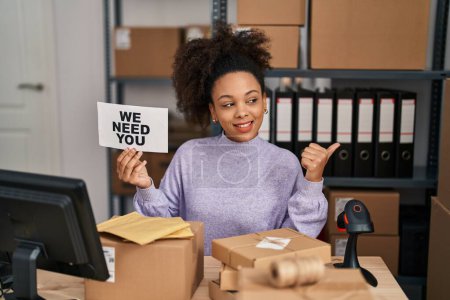 Téléchargez les photos : Jeune femme afro-américaine travaillant dans un commerce électronique de petite entreprise tenant une bannière pointant le pouce vers le côté souriant heureux avec la bouche ouverte - en image libre de droit