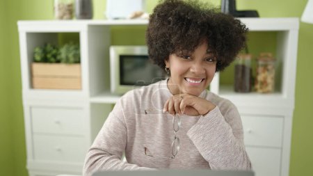 Foto de Joven afroamericana americana sonriendo confiada sentada en la mesa en casa - Imagen libre de derechos