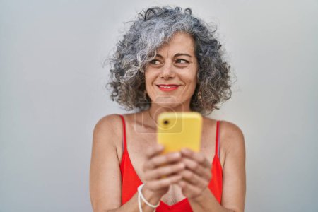 Foto de Mujer de pelo gris de mediana edad sonriendo confiada usando teléfono inteligente sobre fondo aislado blanco - Imagen libre de derechos