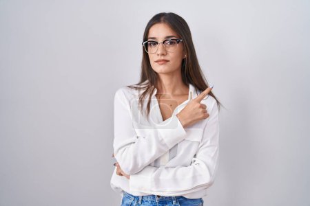 Téléchargez les photos : Jeune femme brune portant des lunettes pointant avec le doigt de la main sur le côté montrant la publicité, visage sérieux et calme - en image libre de droit