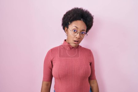 Foto de Hermosa mujer africana con el pelo rizado de pie sobre fondo rosa en la cara de choque, mirando escéptico y sarcástico, sorprendido con la boca abierta - Imagen libre de derechos