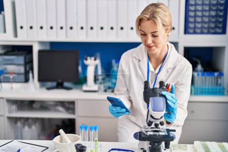 Téléchargez les photos : Jeune femme blonde scientifique utilisant un microscope et un smartphone au laboratoire - en image libre de droit