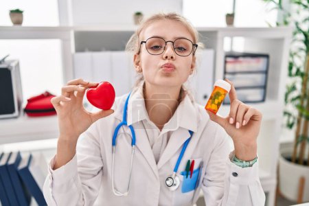 Téléchargez les photos : Jeune femme caucasienne médecin tenant des pilules et le cœur en regardant la caméra souffler un baiser étant belle et sexy. expression d'amour. - en image libre de droit