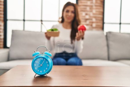 Téléchargez les photos : Jeune femme hispanique tenant salade et pomme assise sur le canapé à la maison - en image libre de droit