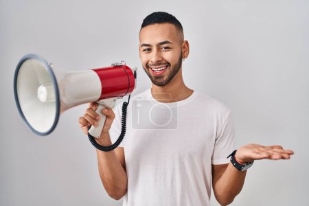 Téléchargez les photos : Jeune homme hispanique criant à travers le mégaphone célébrant la réalisation avec un sourire heureux et l'expression gagnante avec la main levée - en image libre de droit
