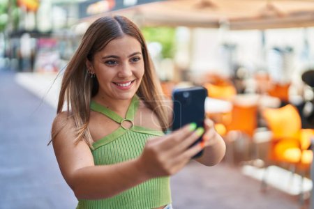 Foto de Joven hermosa mujer hispana sonriendo confiada haciendo selfie por el teléfono inteligente en la terraza de la cafetería - Imagen libre de derechos