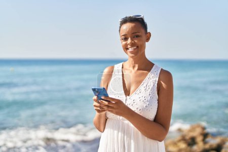 Foto de Mujer afroamericana sonriendo confiada usando smartphone en la playa - Imagen libre de derechos