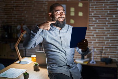 Téléchargez les photos : Jeune homme hispanique avec barbe et tatouages travaillant au bureau la nuit souriant joyeuse montrant et pointant du doigt les dents et la bouche. concept de santé dentaire. - en image libre de droit