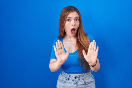 Photo for Redhead woman standing over blue background moving away hands palms showing refusal and denial with afraid and disgusting expression. stop and forbidden. - Royalty Free Image