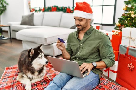 Foto de Joven hispano usando laptop y tarjeta de crédito sentado en el piso con perro por árbol de navidad en casa - Imagen libre de derechos