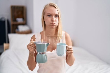 Téléchargez les photos : Jeune femme caucasienne buvant du café sur le lit gonflant les joues avec un visage drôle. bouche gonflée d'air, attrapant l'air. - en image libre de droit