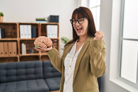 Photo for Young therapist woman working at therapy office holding brain screaming proud, celebrating victory and success very excited with raised arm - Royalty Free Image