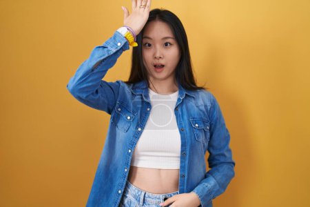 Photo for Young asian woman standing over yellow background surprised with hand on head for mistake, remember error. forgot, bad memory concept. - Royalty Free Image