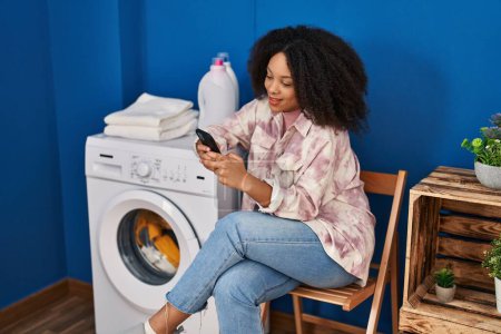 Téléchargez les photos : Jeune femme afro-américaine utilisant un smartphone attendant la machine à laver à la blanchisserie - en image libre de droit