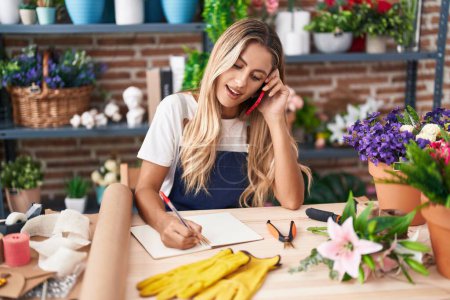 Photo for Young blonde woman florist talking on smartphone writing on notebook at florist - Royalty Free Image