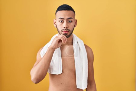 Photo for Young hispanic man standing shirtless with towel with hand on chin thinking about question, pensive expression. smiling with thoughtful face. doubt concept. - Royalty Free Image
