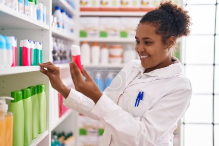 Foto de Farmacéutica afroamericana sonriendo confiada sosteniendo pasta de dientes de estanterías en la farmacia - Imagen libre de derechos