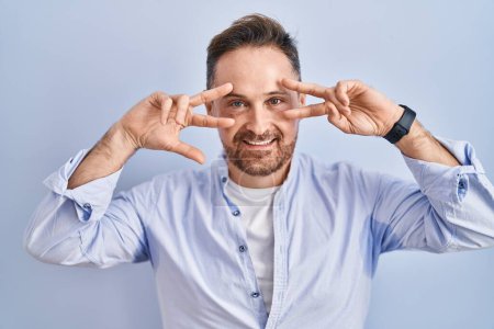 Photo for Middle age caucasian man standing over blue background doing peace symbol with fingers over face, smiling cheerful showing victory - Royalty Free Image
