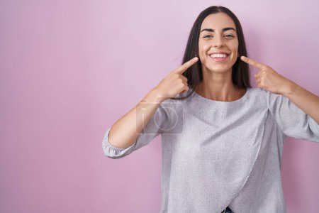 Téléchargez les photos : Jeune femme brune debout sur fond rose souriant gai montrant et pointant avec les doigts dents et la bouche. concept de santé dentaire. - en image libre de droit