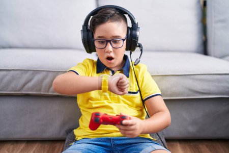 Téléchargez les photos : Jeune enfant hispanique jouant contrôleur de tenue de jeu vidéo portant des écouteurs regardant le temps de la montre inquiet, peur de se faire tard - en image libre de droit