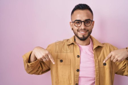 Foto de Joven hombre hispano de pie sobre fondo rosa mirando confiado con sonrisa en la cara, señalándose con los dedos orgullosos y felices. - Imagen libre de derechos